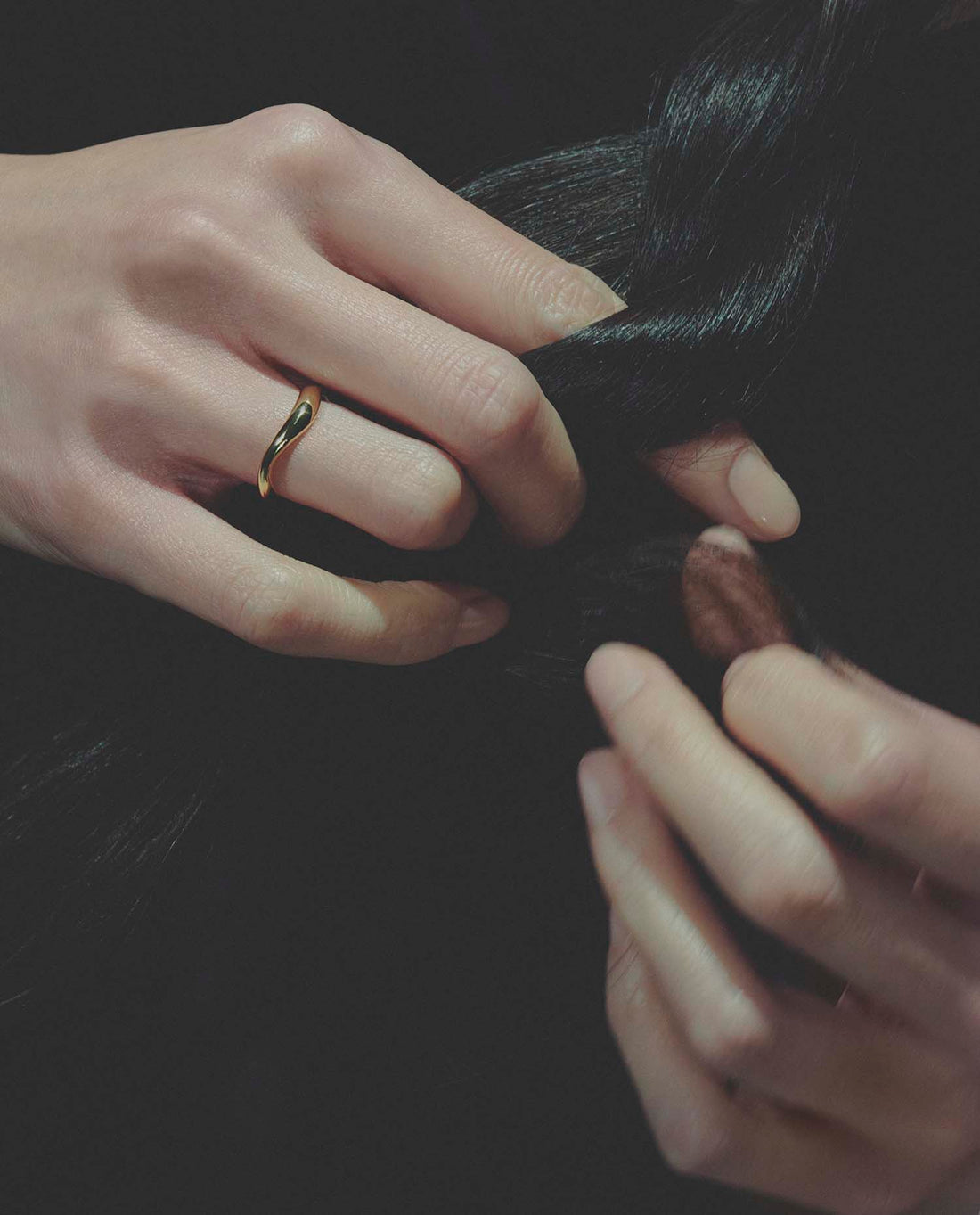 Close-up of hands, wearing a gold ring, braiding hair with a dark background