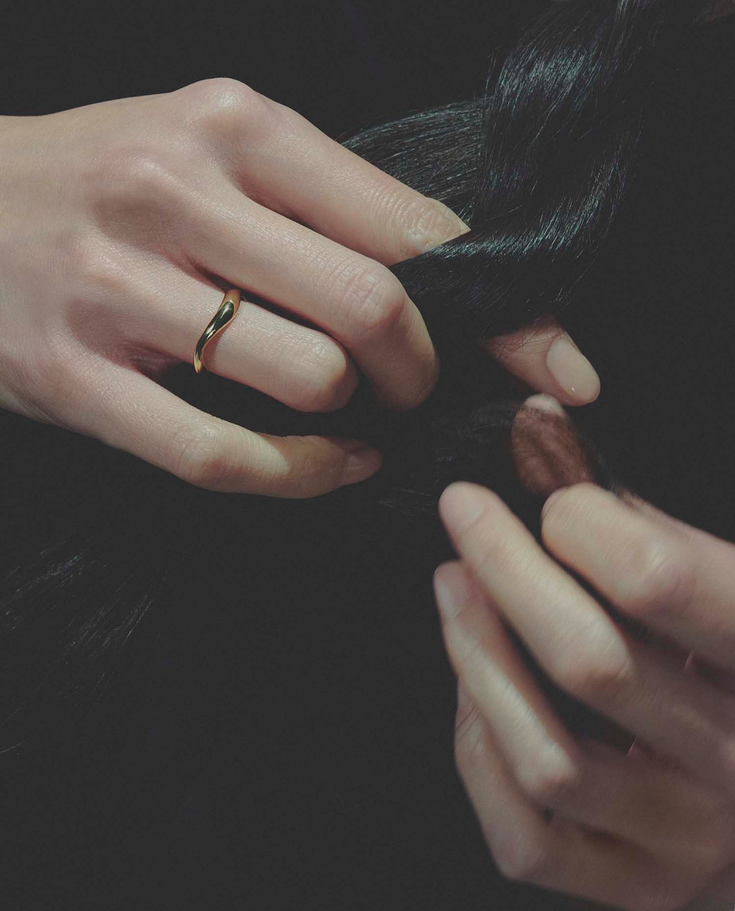 Close-up of hands, wearing a gold ring, braiding hair with a dark background