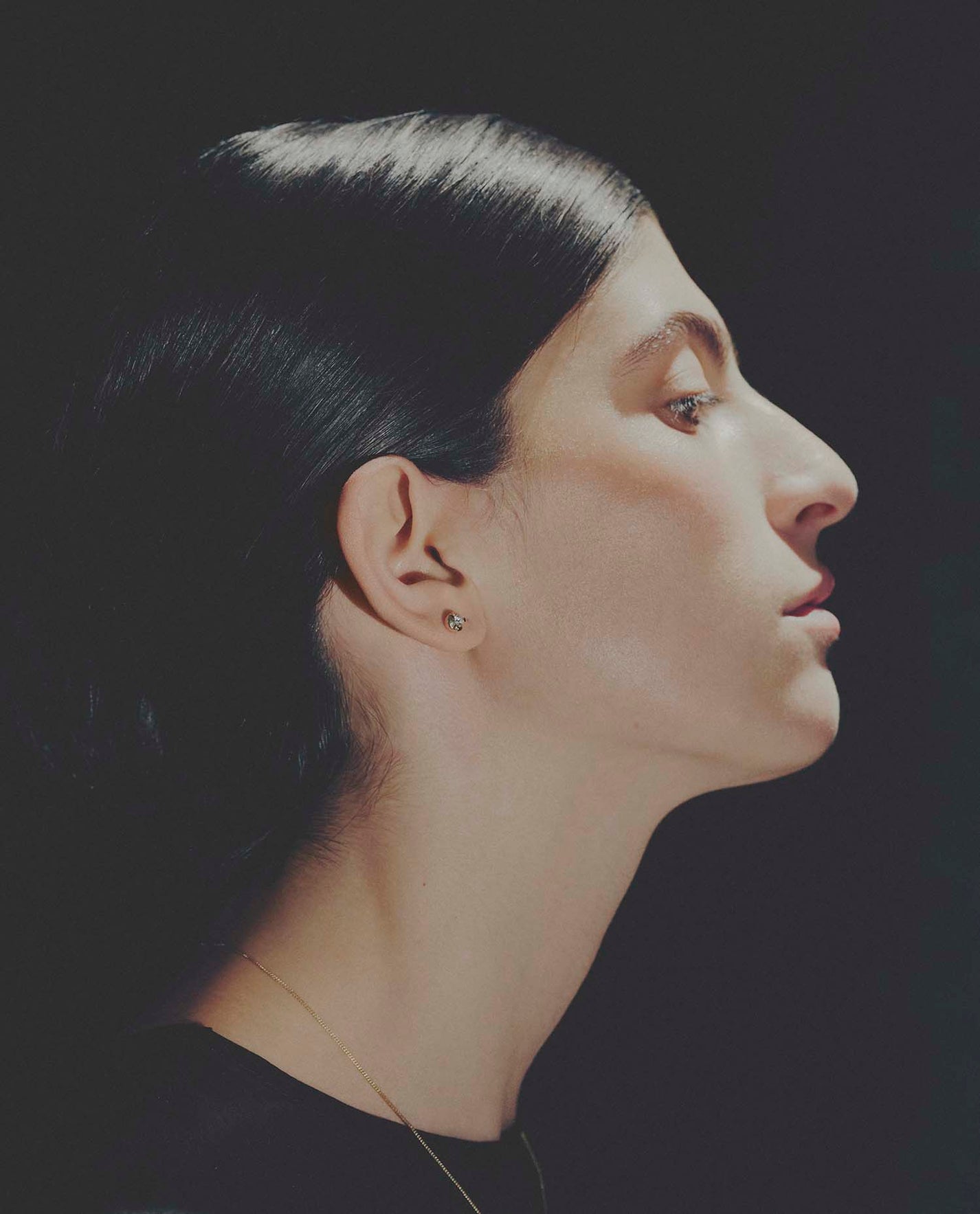 Side profile of a person with dark hair wearing a diamond earring against a black background