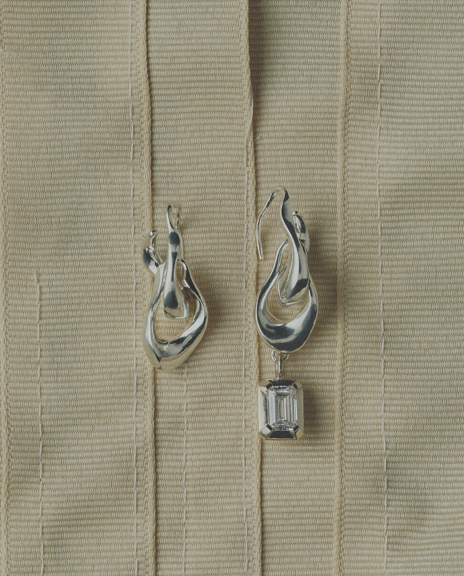 Pair of silver earrings one featuring a diamond on a beige ribbon background