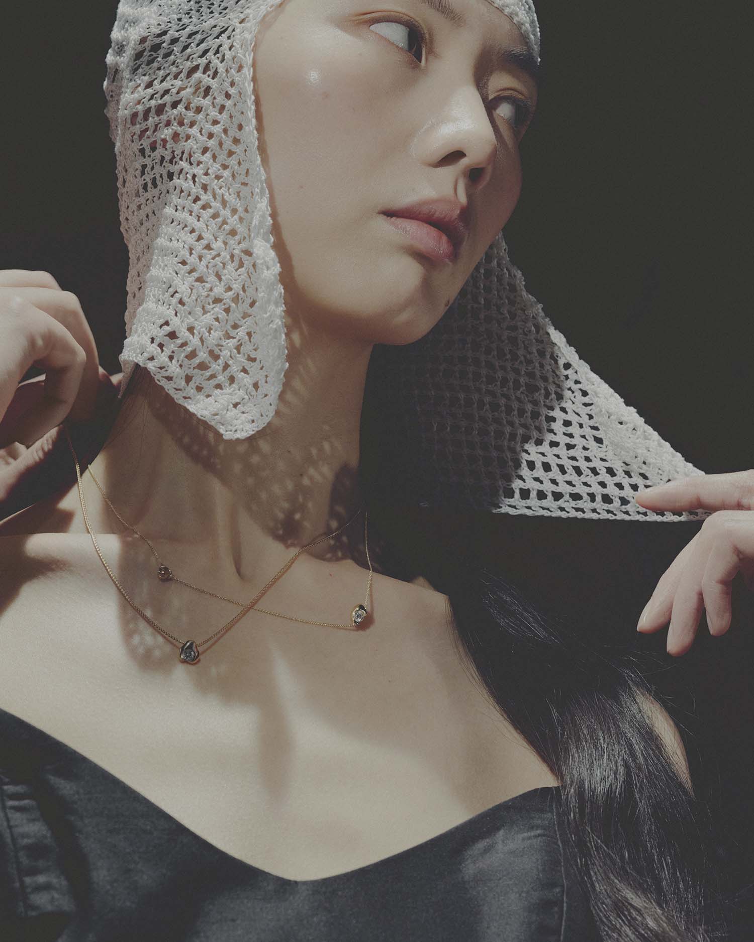 Woman wearing two gold neckalces, a fishnet head covering and black top against a dark background