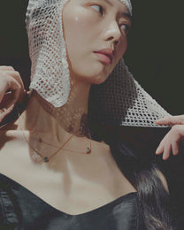 Woman wearing two gold neckalces, a fishnet head covering and black top against a dark background